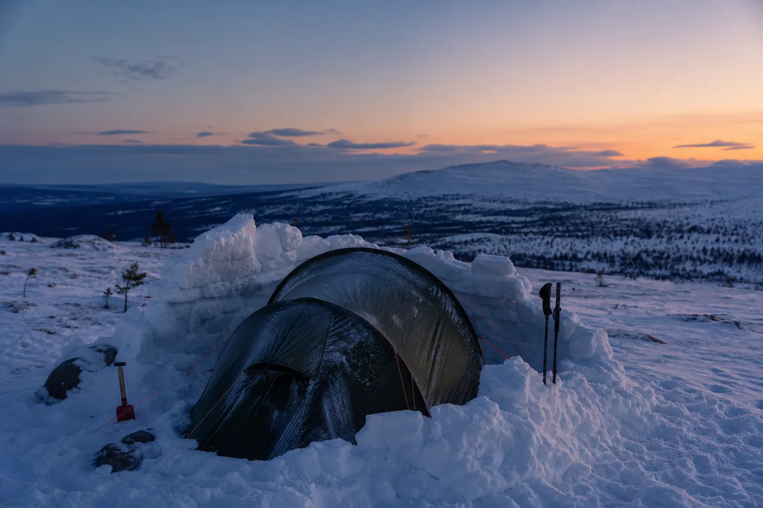 Snötäckt kupoltält med snömur på vindsidan i fjällterräng under vinterkväll