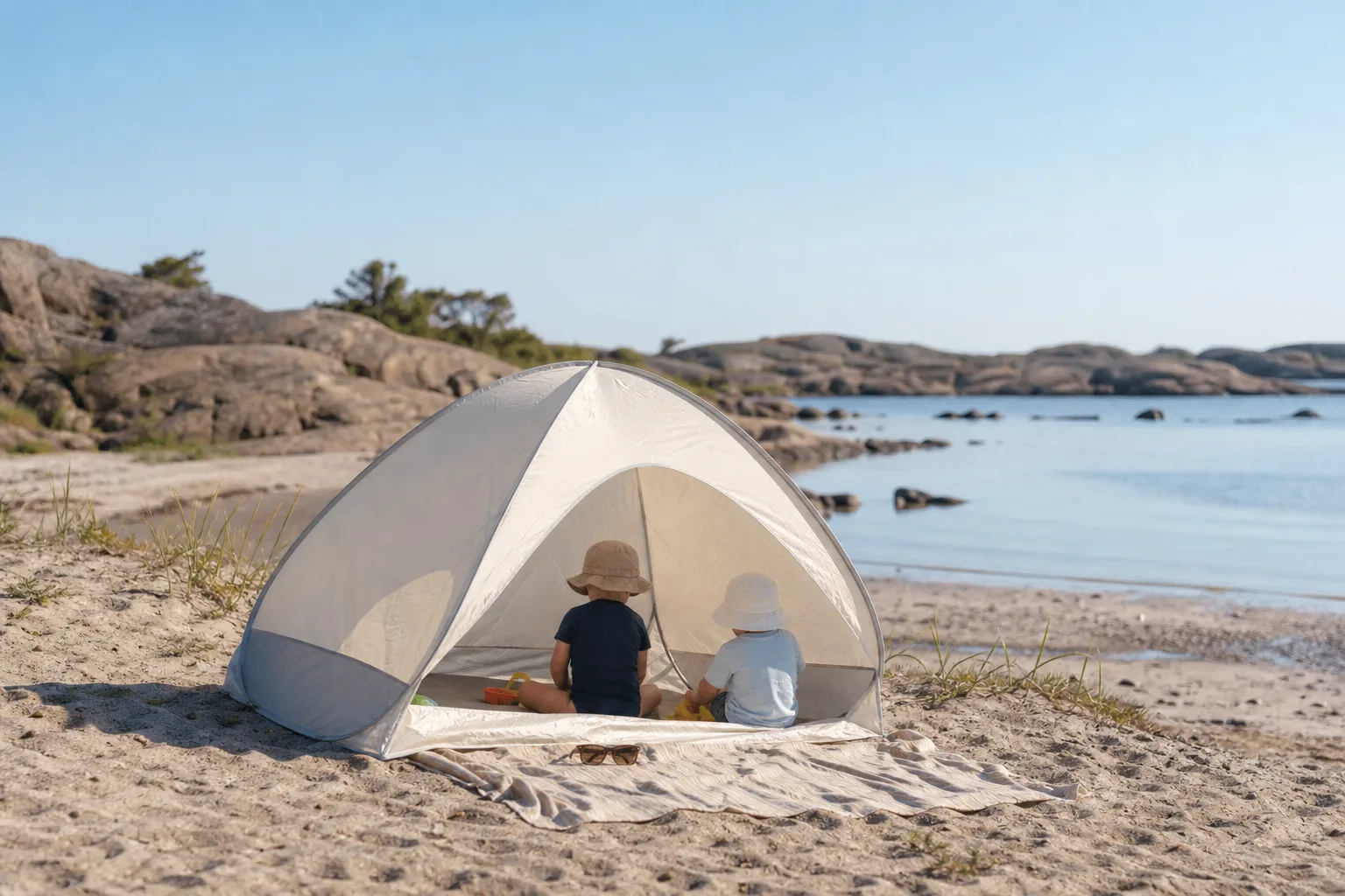 Litet UV-tält i ljust tyg uppfällt på en sandstrand med solglasögon och handduk bredvid
